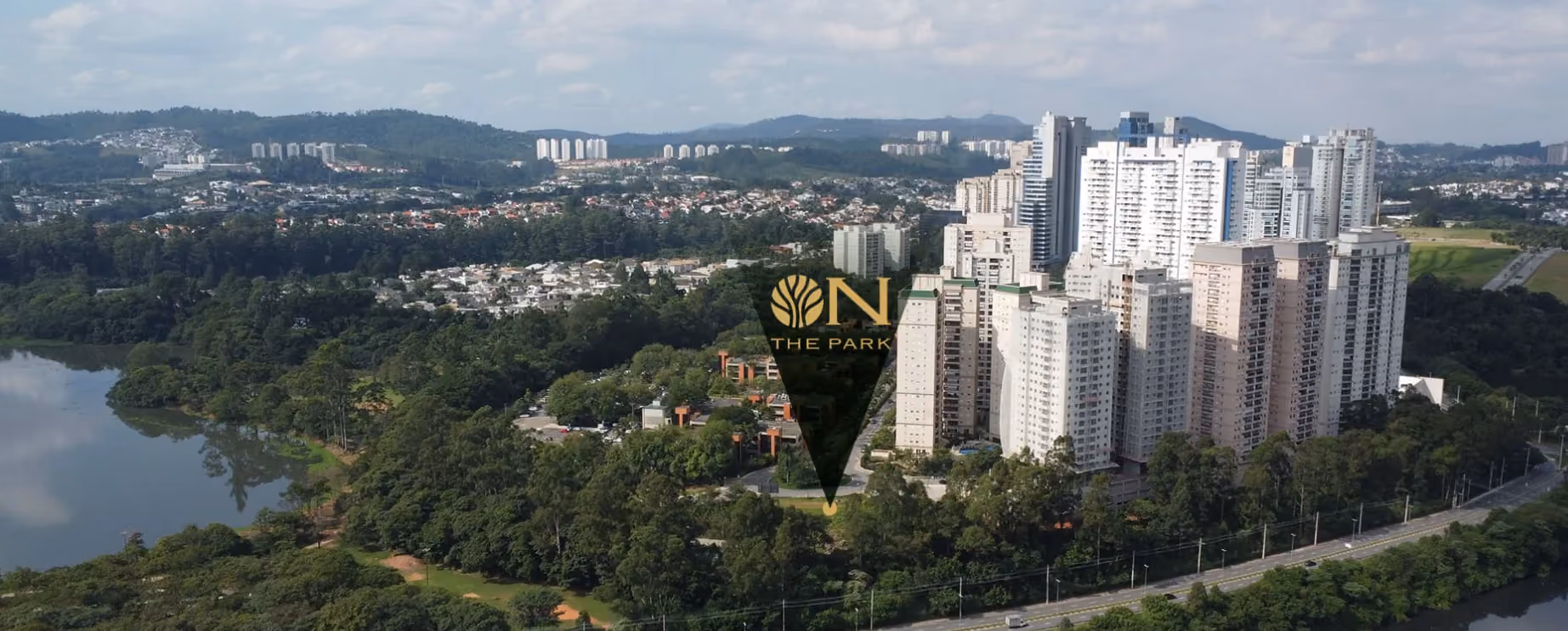 Vista aérea antes da construção, com água à esquerda, a mata, condomínios de prédios claros à direita, uma avenida em primeiro plano e um marcador com o logo On The Park sobre o terreno