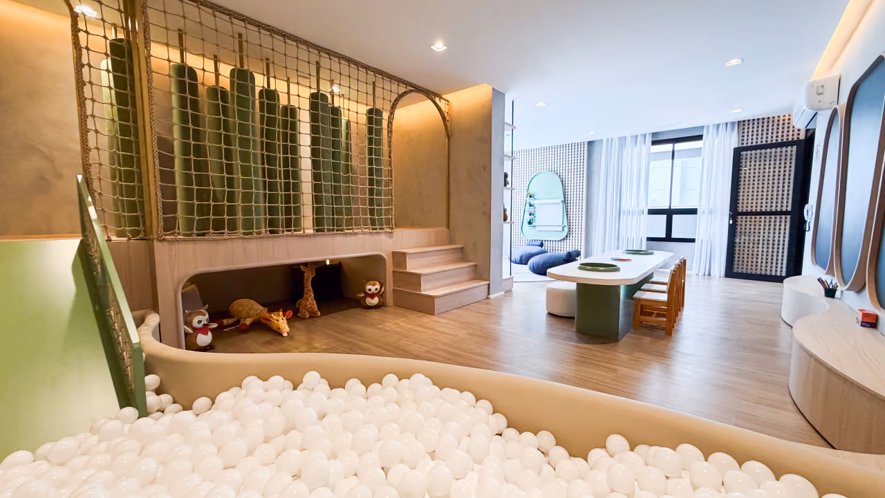 Brinquedoteca com piscina de bolinhas brancas, rede de escalada com rolos verdes, bichos de pelúcia, mesa infantil com cadeirinhas e janelas com cortinas claras