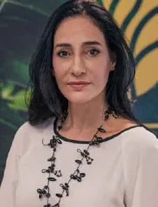 Retrato de Camila Pupo, arquiteta de cabelos pretos longos, com blusa clara e colar preto, diante de painel verde e dourado