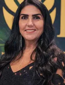 Retrato de Claudia Albertini, arquiteta de cabelos pretos longos e ondulados, sorrindo, com blusa preta rendada e brincos longos, diante de painel verde e dourado