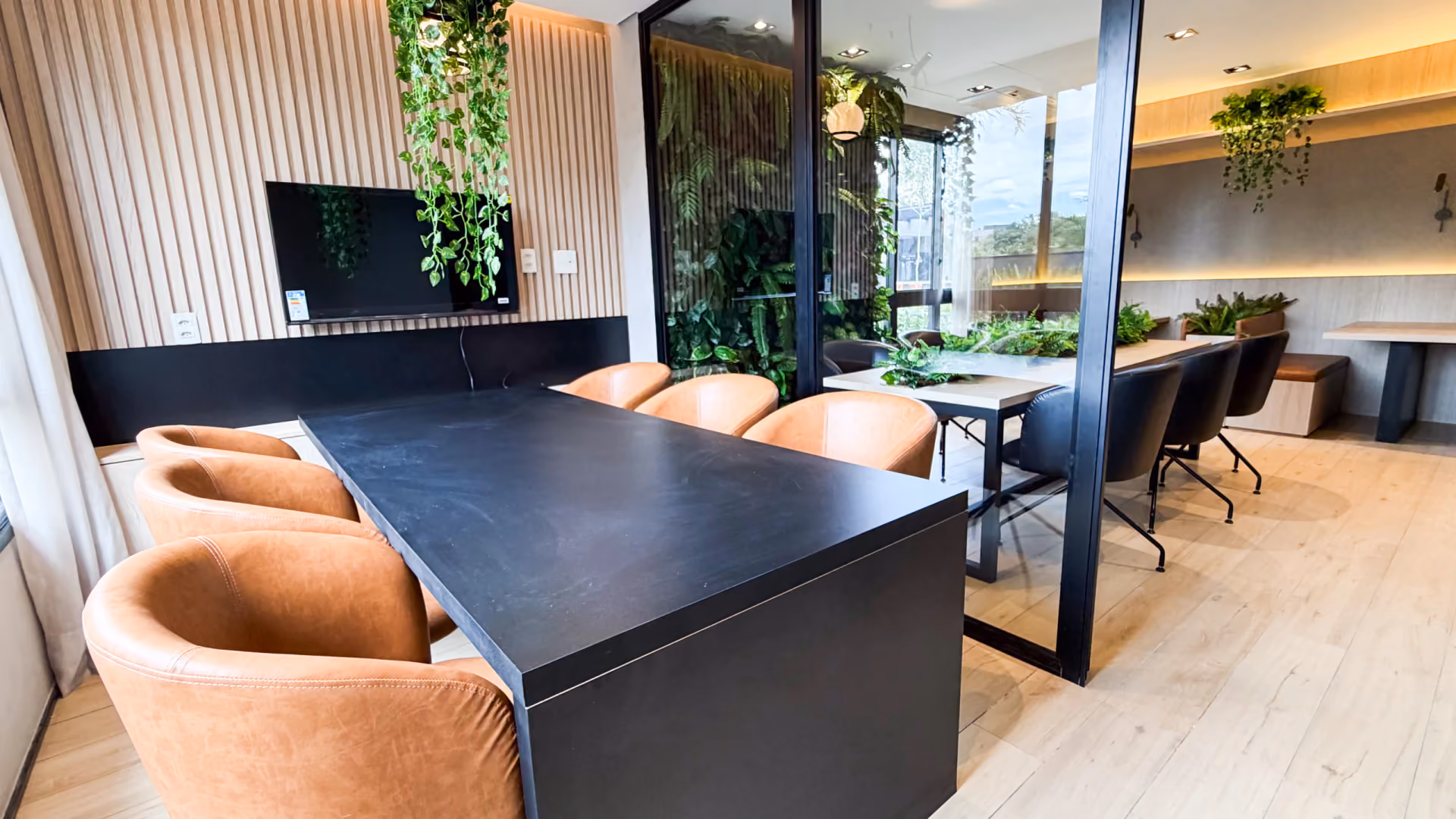 Coworking com mesa escura, poltronas de couro caramelo, televisão em painel ripado e divisória de vidro para a sala com jardim vertical
