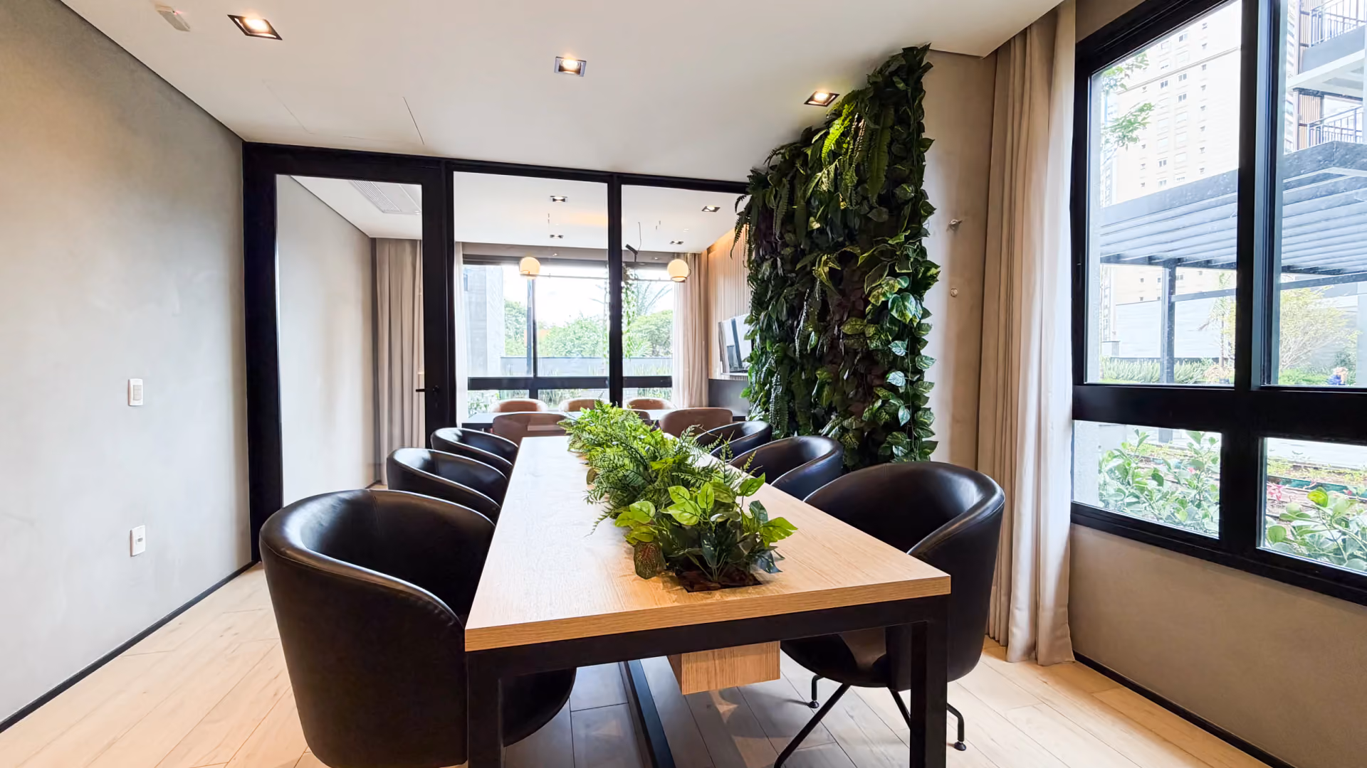 Sala de reunião do coworking com mesa longa de madeira com plantas no centro, poltronas de couro preto, jardim vertical e janela para o jardim