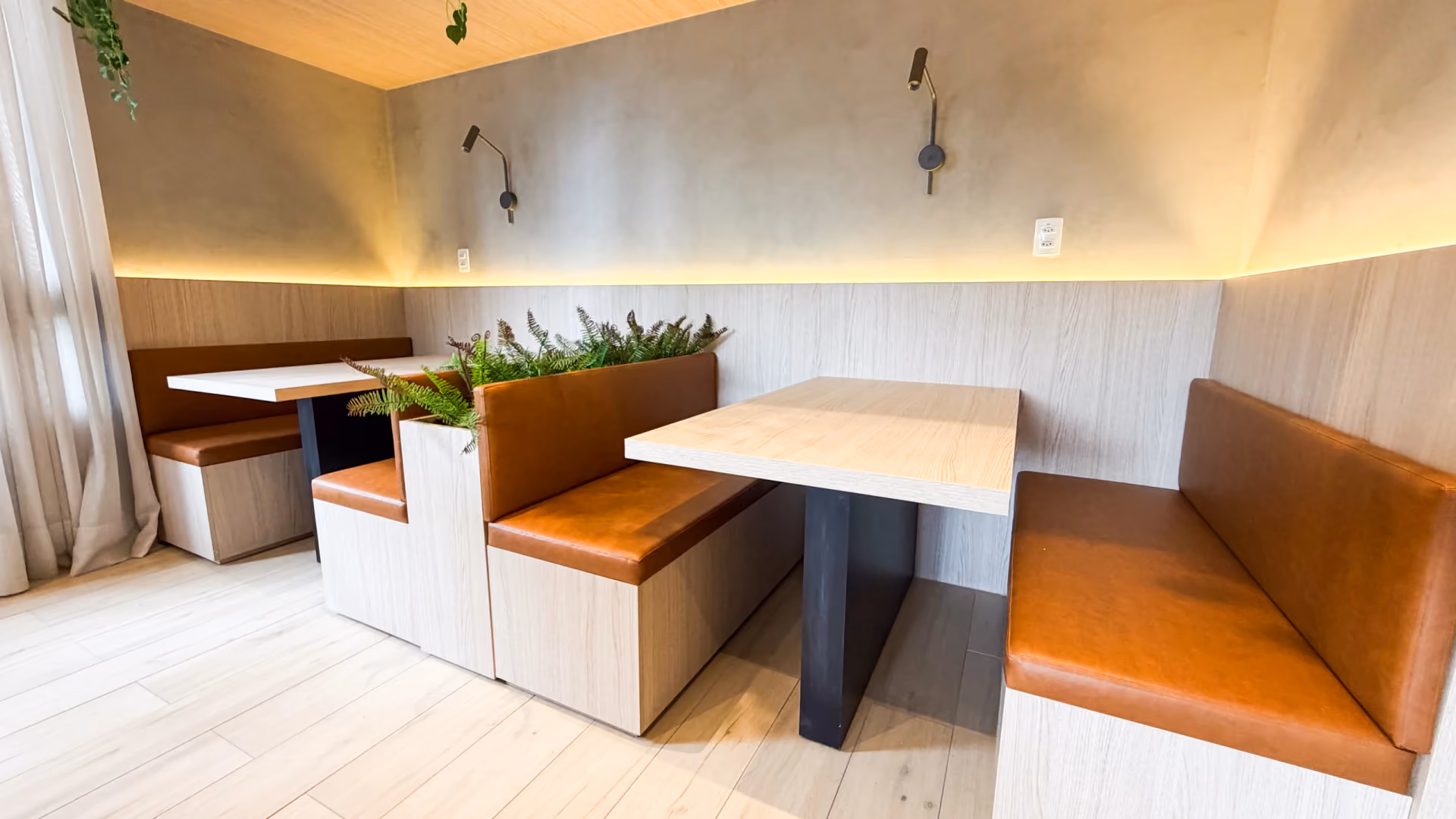 Cabines do coworking com bancos estofados caramelo, mesas de madeira clara, floreira e arandelas com iluminação indireta