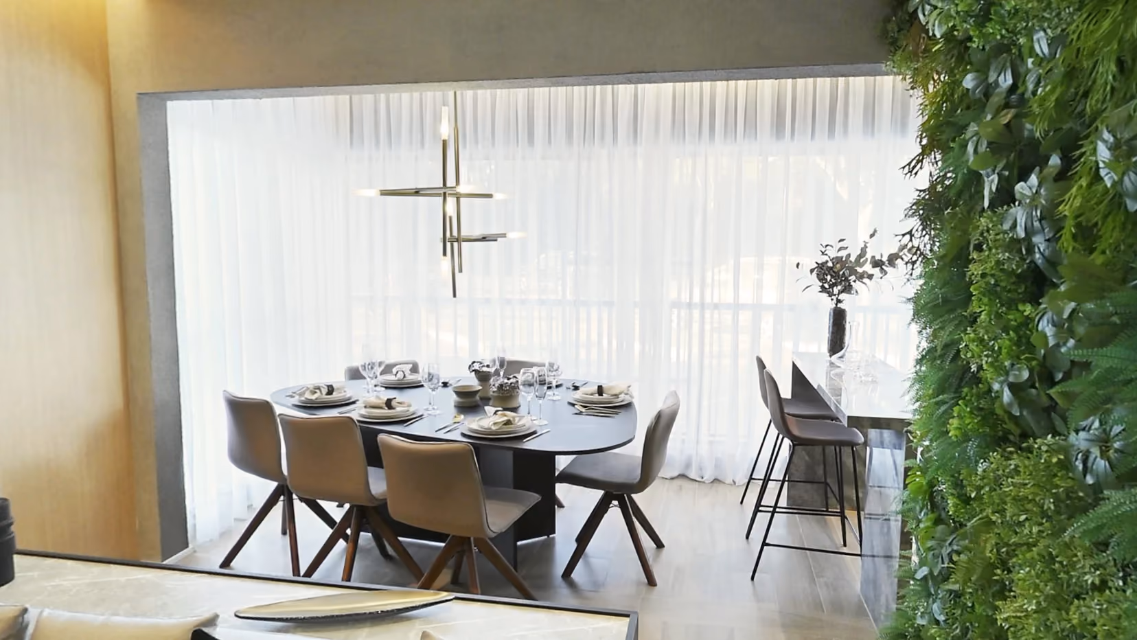 Sala de jantar do apartamento decorado com mesa oval posta para seis, cadeiras de couro, lustre de hastes douradas e cortina clara na varanda, e parede verde de plantas à direita