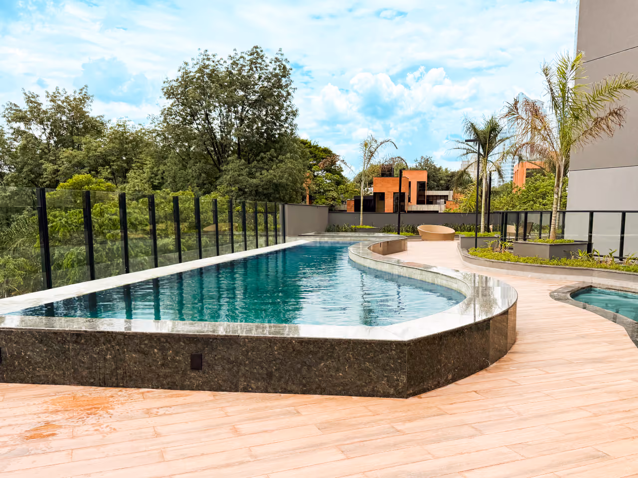 Piscina adulto de borda curva revestida de granito escuro, deck amadeirado, guarda-corpo de vidro, palmeiras e mata ao fundo, e a borda da piscina infantil à direita