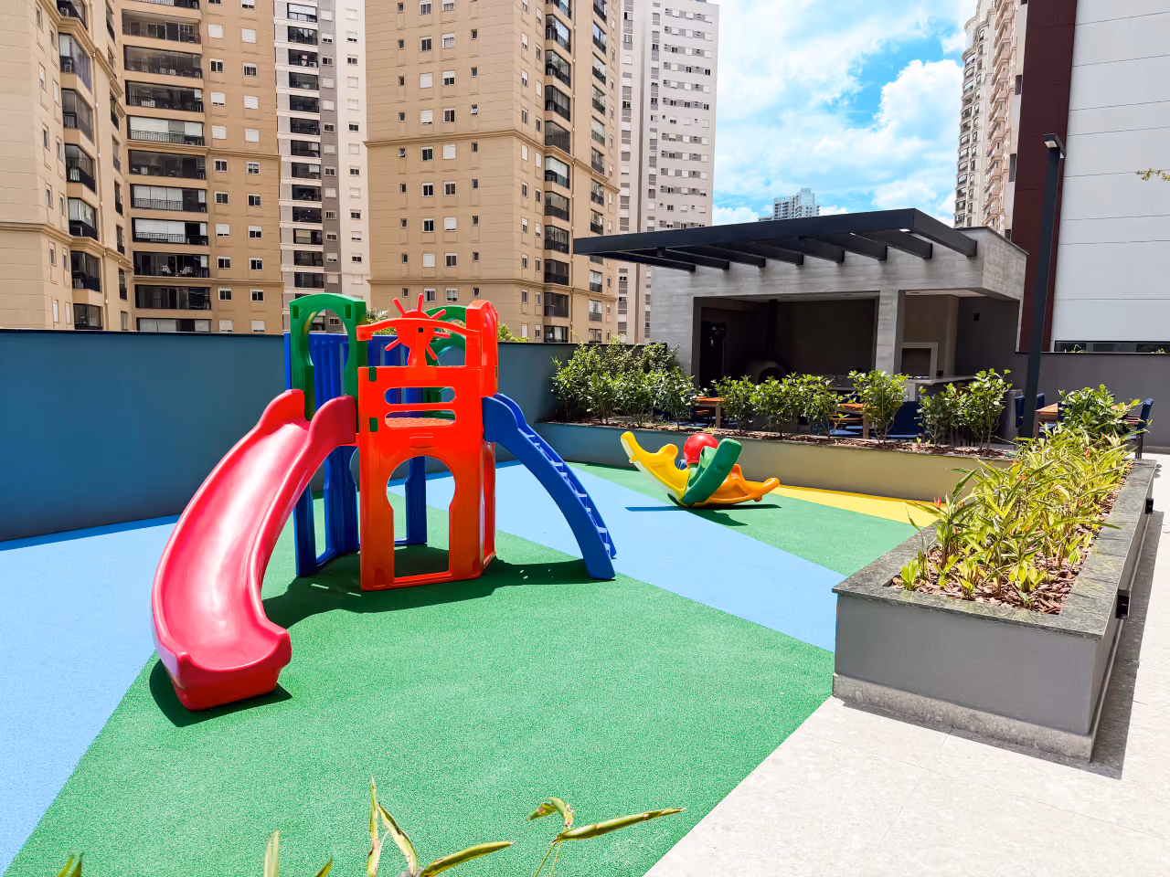 Playground com piso emborrachado em faixas verdes, azuis e amarelas, brinquedo com escorregadores vermelho e azul, gangorra, canteiros e o quiosque da churrasqueira ao fundo, entre prédios