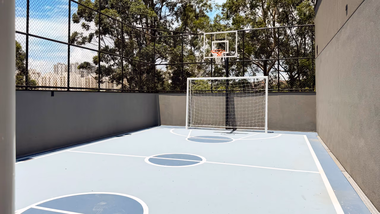 Quadra recreativa com piso azul e linhas brancas, gol, tabela de basquete e alambrado alto com árvores ao fundo