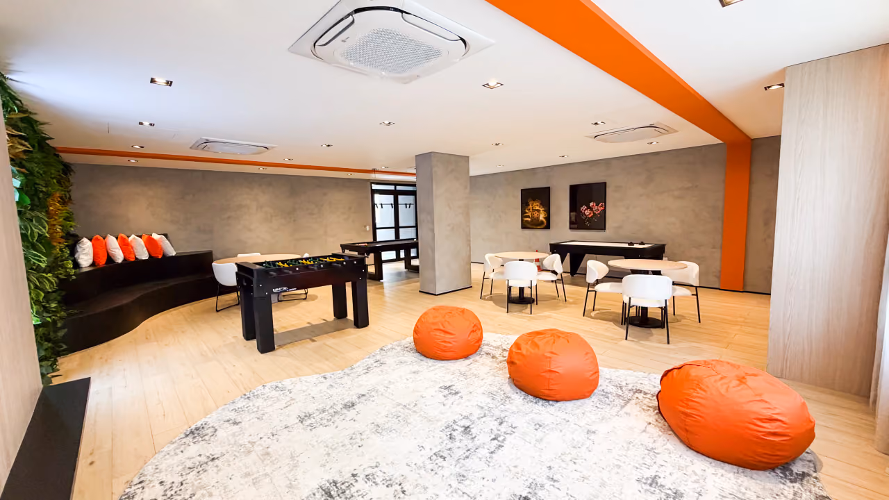 Salão de jogos com pufes laranja sobre tapete, pebolim, air hockey, mesas redondas com cadeiras brancas, sofá escuro com almofadas e jardim vertical