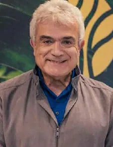 Retrato de Sergio Santana, paisagista de cabelos grisalhos, sorrindo, com jaqueta cinza sobre camisa azul, diante de painel verde e dourado